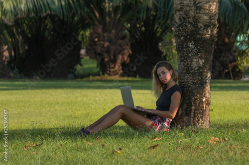 young blonde sits under a palm tree in green park, student preparing for classes