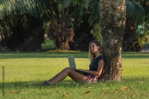 young beautiful woman in black top with laptop on her knees sits under palm