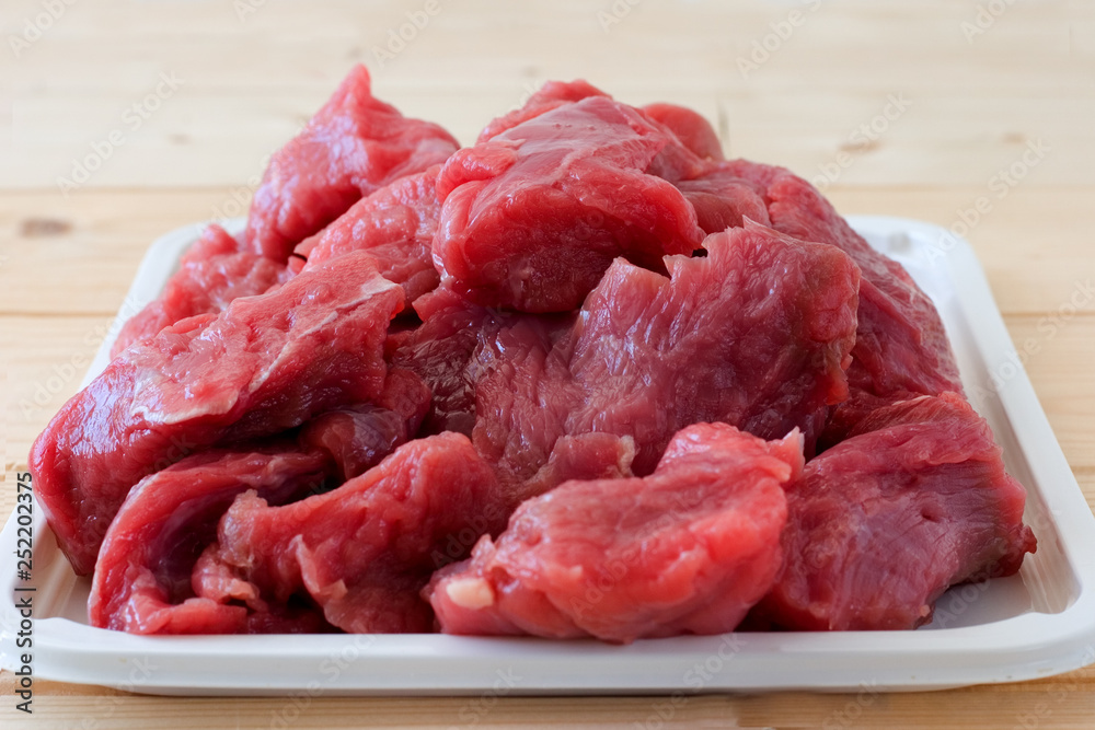 Raw fresh beef meat pieces on a white tray on a a beige plank wooden table. Close up Raw fresh beef meat pieces on a white tray on a a beige plank wooden table. Close up