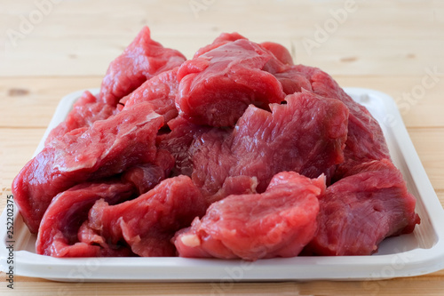 Raw fresh beef meat pieces on a white tray on a a beige plank wooden table. Close up