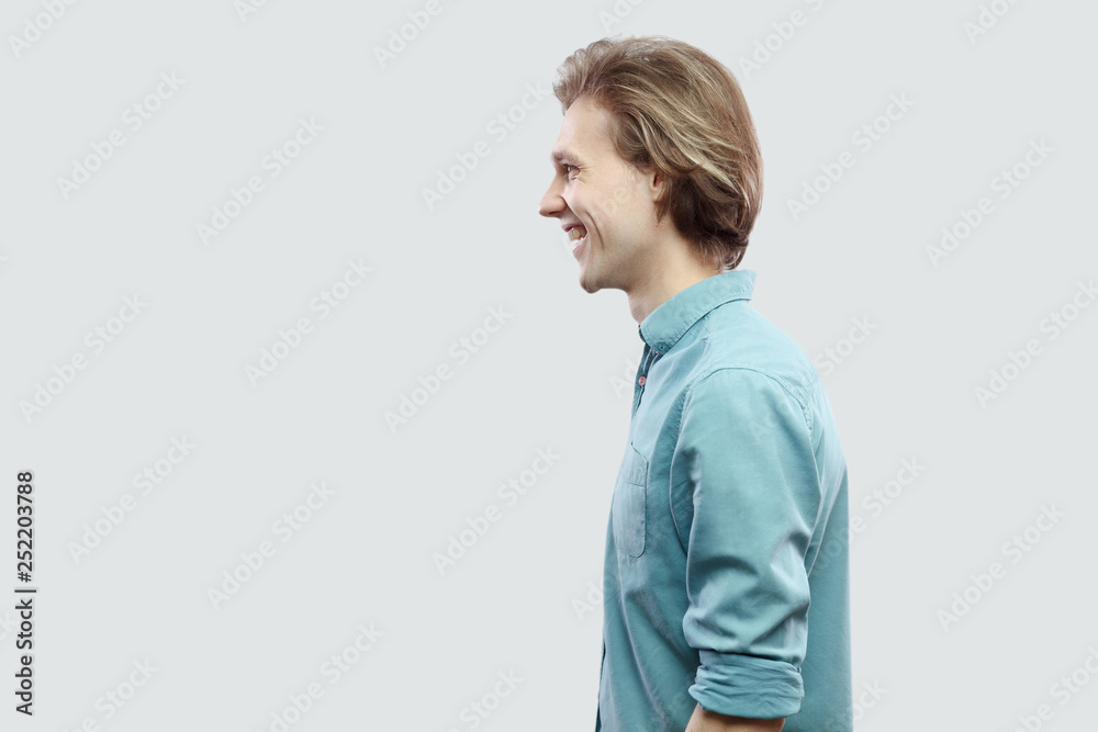 Profile side view portrait of happy handsome long haired blonde young man in blue casual shirt standing and looking with toothy smile. indoor studio shot, isolated on light grey background. Profile side view portrait of happy handsome long haired blonde young man in blue casual shirt standing and looking with toothy smile. indoor studio shot, isolated on light grey background.