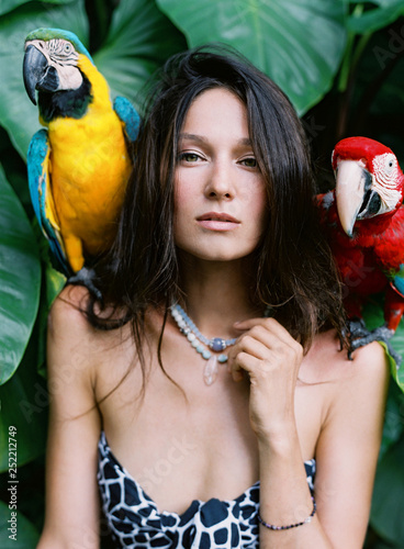 Portrait of young attractive brunette woman with ara parrots on her shoulders on tropical background.