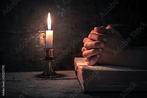 Close up hands of man praying on bible with light of candle on wooden table at worship room. christian concept.