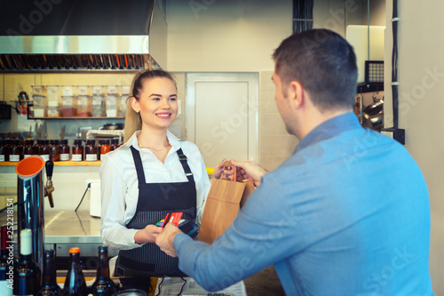 Customer using credit card to pay order on reader with nfc technology holded by smiling waitress working at shop counter. 