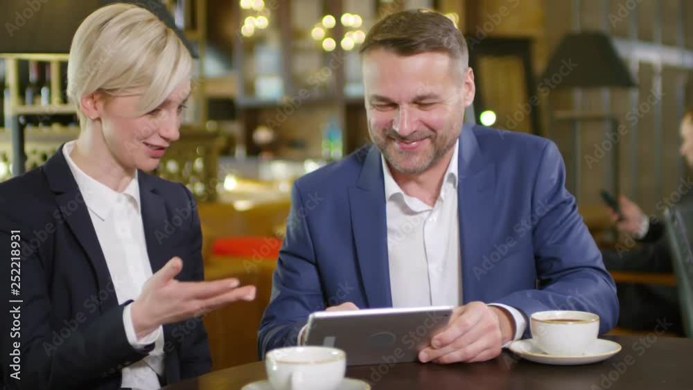 Medium shot of male and female business partners sitting in cafe during coffee break, looking at tablet screen and discussing news with smile on their faces Medium shot of male and female business partners sitting in cafe during coffee break, looking at tablet screen and discussing news with smile on their faces