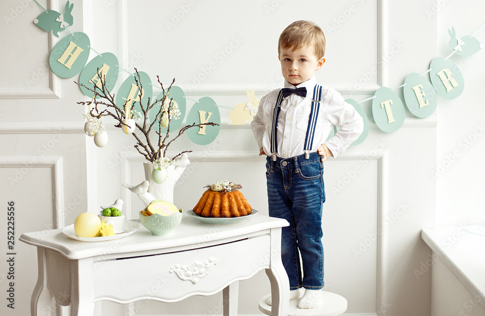 Cute boy in a white shirt and tassel butterfly stands on a white chair near a table in a white room on a table. Easter decoration of a vase with branches and Easter eggs of gingerbread rabbits. Cute boy in a white shirt and tassel butterfly stands on a white chair near a table in a white room on a table. Easter decoration of a vase with branches and Easter eggs of gingerbread rabbits.