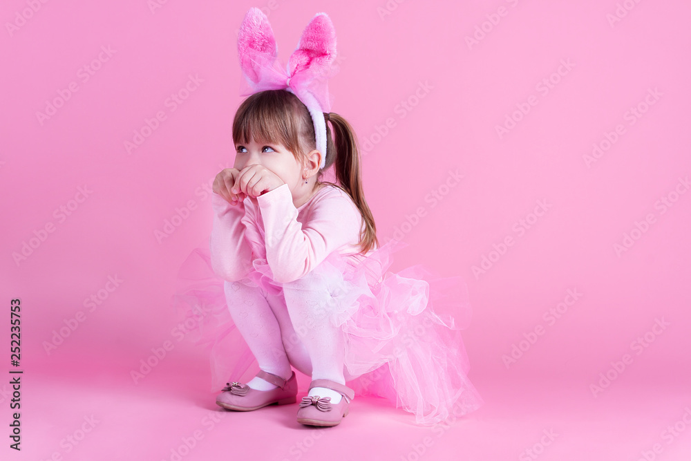 Cute pink young girl child daughter wears pink dress like rabbit playing in easter holiday game with rabbit ears on isolated pink background. Spring is coming and Easter holiday concept