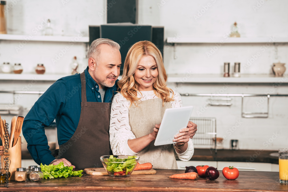 happy woman using digital tablet while standing with husband in kitchen near vegetables happy woman using digital tablet while standing with husband in kitchen near vegetables