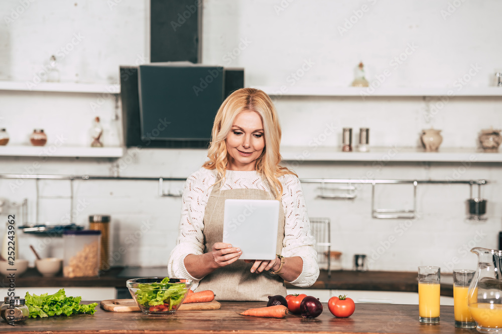happy woman using digital tablet while standing in kitchen near vegetables happy woman using digital tablet while standing in kitchen near vegetables