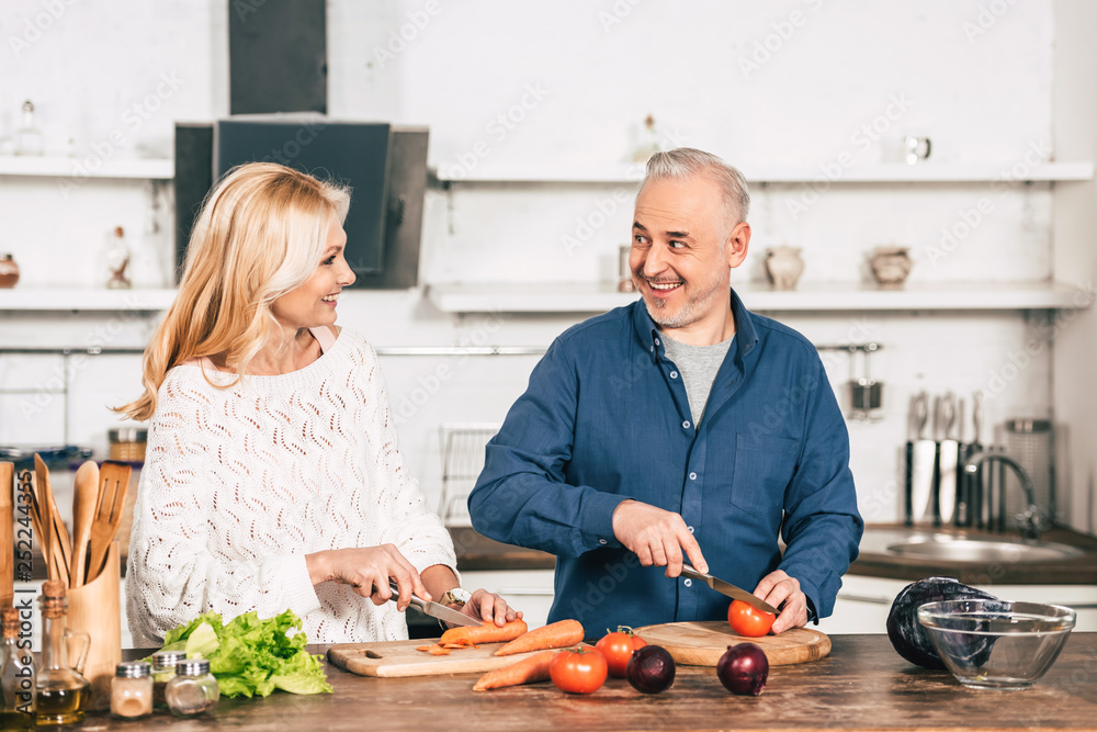 cheerful couple cutting carrot and tomato and smiling in kitchen