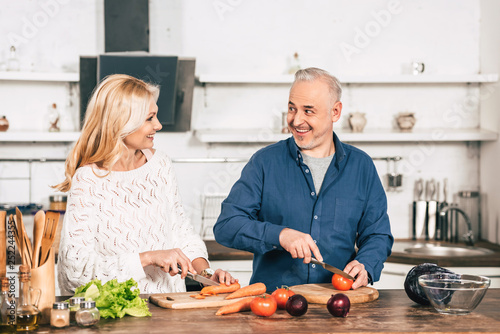 cheerful couple cutting carrot and tomato and smiling in kitchen