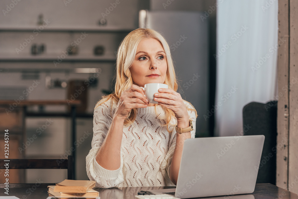 attractive pensive woman holding cup with drink near laptop attractive pensive woman holding cup with drink near laptop