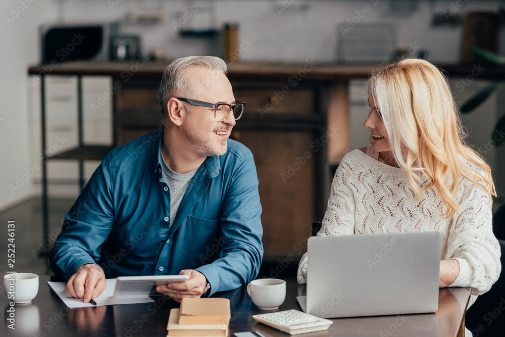 handsome man in glasses using digital tablet and looking at attractive blonde wife with laptop handsome man in glasses using digital tablet and looking at attractive blonde wife with laptop