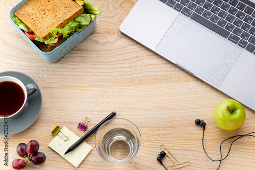 Overhead image of healthy sandwich in plastic container with some fruits on wooden table next to opened laptop. Daily office or school lunch concept