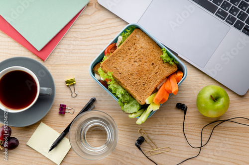 Quadro su tela Overhead image of healthy sandwich in plastic container with some fruits on wooden table next to opened laptop