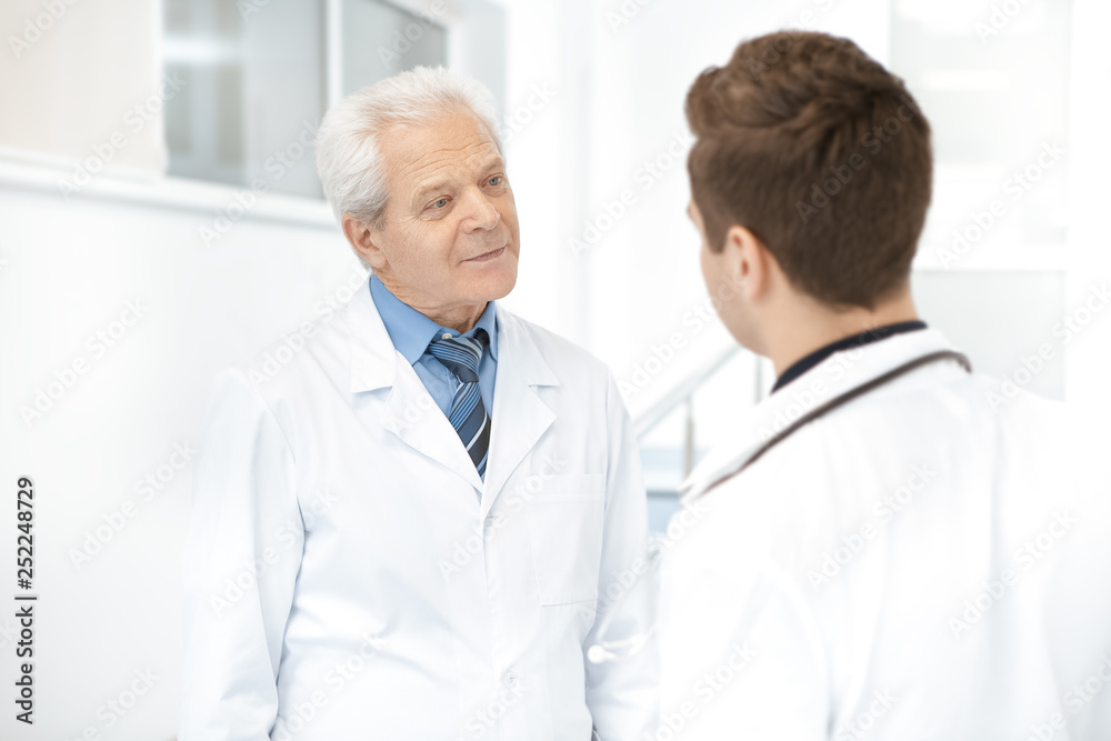 Elderly doctor having a discussion with his younger colleague at the hospital hallway Elderly doctor having a discussion with his younger colleague at the hospital hallway