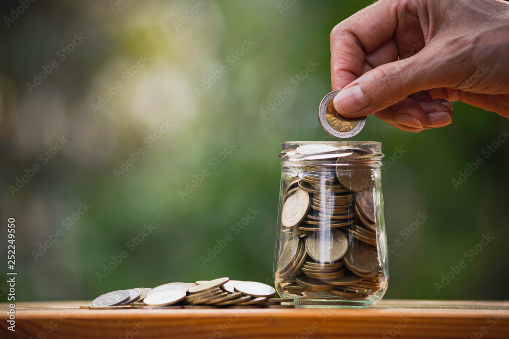 Hand of person holding a coin putting in glass jar on wooden in nature background to save money for the future plan. Hand of person holding a coin putting in glass jar on wooden in nature background to save money for the future plan.