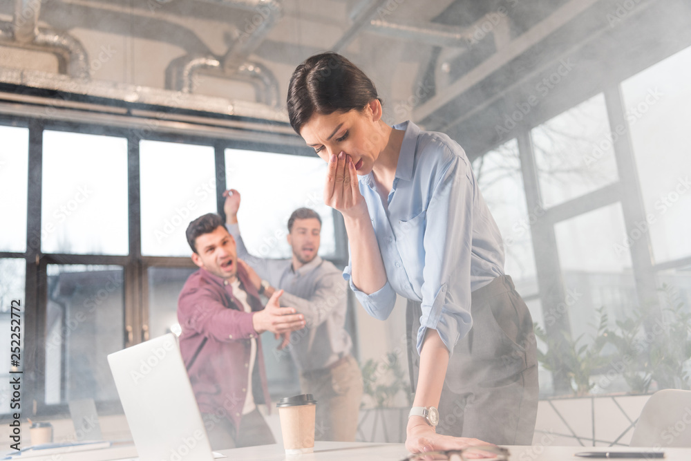 businesswoman holding nose while standing near coworkers in office with smoke