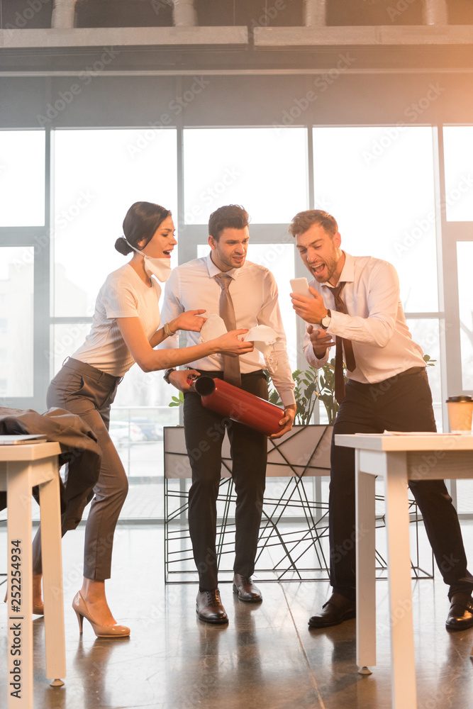 Obraz premium terrified businessman holding extinguisher near yelling coworkers looking at smartphone in office