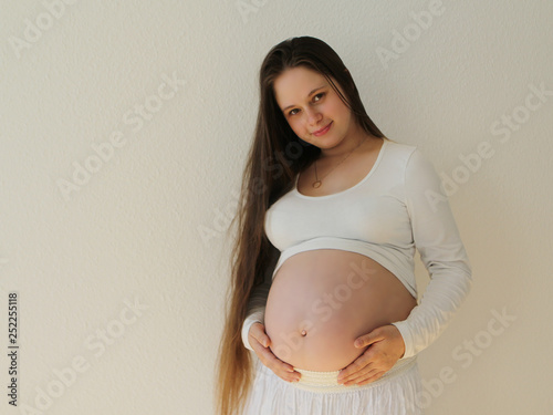 Pregnant woman in white clothes on a white background gently supports the stomach with open hands in the room at home. Light comes from a window on a white background wallpaper. Close-up.