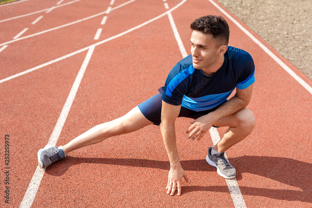 Obraz premium Marathon preparation. Young man on stadium outside stretching on track smiling happy