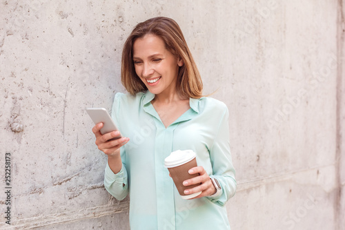 Young woman standing isolated leaning on wall drinking coffee checking social media on smartphone