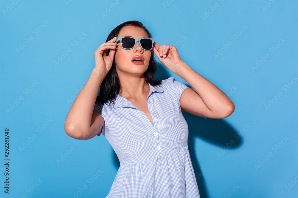 Freestyle. Woman in dress wearing eyeglasses standing isolated on blue wall smiling glamorous