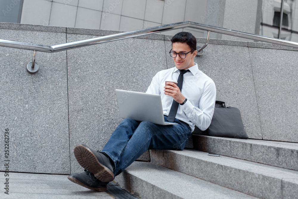 Morning routine. Businessman in eyeglasses sitting on stairs on the city street drinking hot coffee checking mail on laptop joyful Morning routine. Businessman in eyeglasses sitting on stairs on the city street drinking hot coffee checking mail on laptop joyful