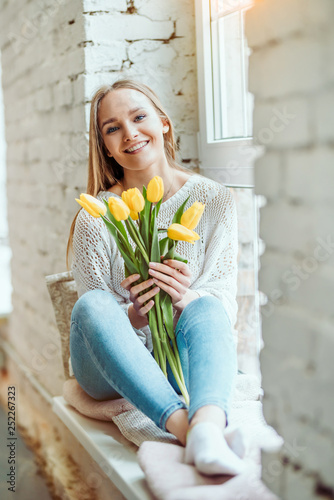 Portrait of beautiful woman with bouquet of yellow tulips sitting on the window sill and looking at camera. © HBS