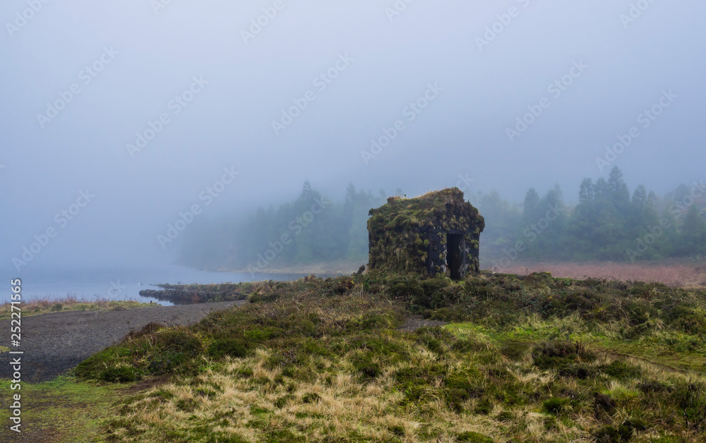 little stone hut covered by moss at the shallow banks of lake Lagoa Rasa surrounded by green forest lost in mist. mysterious moody day, the island of Sao Miguel, Azores, Portugal