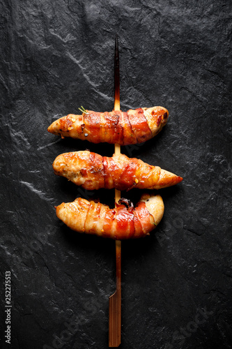 Grilled skewers of chicken tenderloin wrapped with bacon on a black background, top view