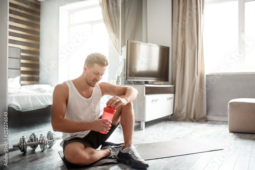 Young well-built man go in for sports in apartment. Nice guy sit on carimate and hold red bottle with water. Workout break. Smile. photo