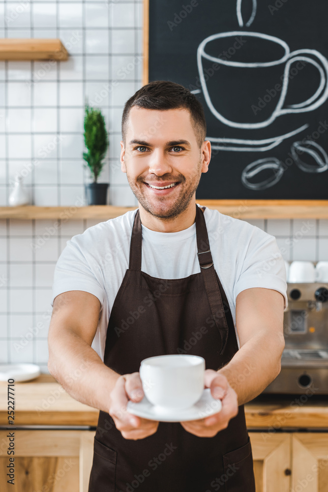 selective focus of handsome barista in brown apron holding cup and saucer in coffee house selective focus of handsome barista in brown apron holding cup and saucer in coffee house