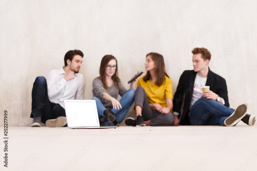Team of positive successful managers of businessmen and designers work together sitting on the floor in creative studio office. Laptop in focus on background blurred student