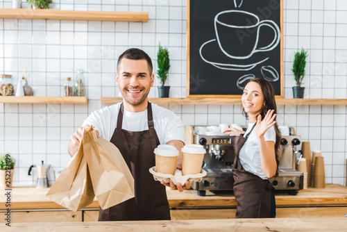 handsome cashier holding paper cups and bags wile attractive brunette barista wavingin in coffee house photo