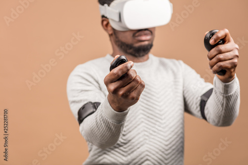Black guy with virtual reality headset playing virtual Race game , moving hands with joysticks like a professional driver in street races , isolated on beige © alfa27