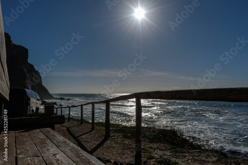 Wooden benches and fence with car parked next to cliff on Porto Barril beach with bright sun, Ericeira - Mafra, Portugal