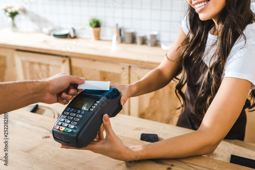 cropped view of cashier standing near bar counter in brown apron and holding terminal wile man paying with credit card in coffee house