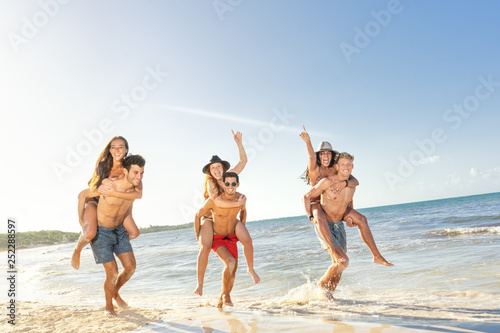 Happy group of young friends doing piggyback ride at the beach. Dutch angle
