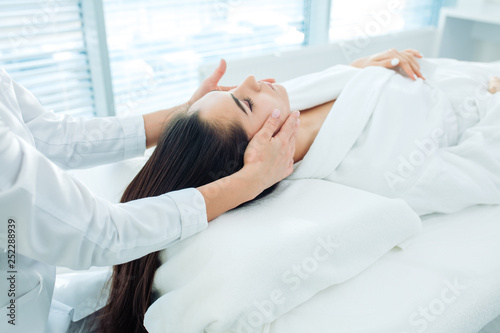Facial spa cosmetology procedure, top view. Skin care lift anti age massage. Caucasian brunette woman dressed in white bathrobe, receiving anti-wrinkle and skin rejuvenation treatment at beauty salon.