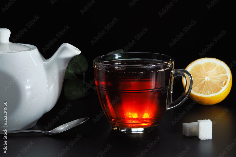Tea is poured into a transparent cup of white teapot, white sugar and lemon on a black background. Tea is poured into a transparent cup of white teapot, white sugar and lemon on a black background.