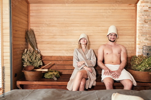 Foto Young couple of tourist relaxing in a sauna sitting on wooden beach with bath bowls and brooms