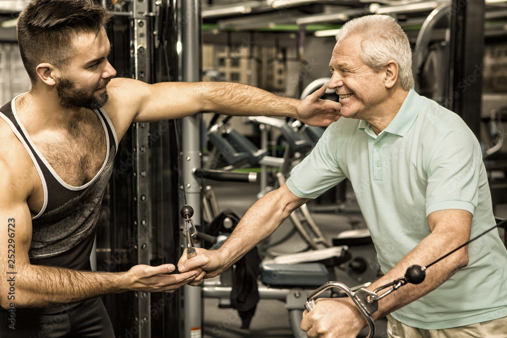 Senior man exercising on cable crossover machine at the gym with a personal trainer Senior man exercising on cable crossover machine at the gym with a personal trainer