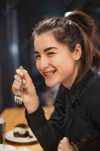 Gorgeous smiling young woman with dark hair eating cake and drinking coffee at a cafeteria in the evening sitting by the window. selective focus  noise effect