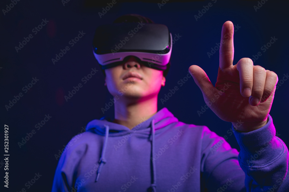 Young Asian guy touching virtual objects during watching 3 D film using virtual reality headset, posing isolated over dark violet background.