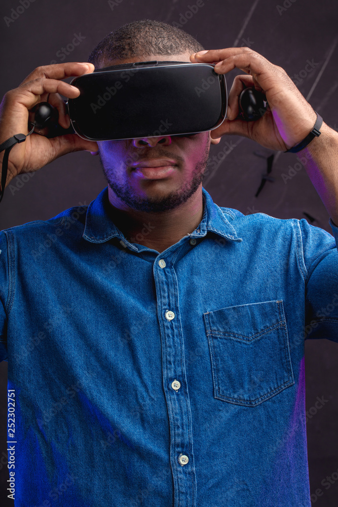 Handsome african designer in denim wearing VR glasses while working over virtual project, posing isolated against dark wall with copy space. Smart innovative device for work and entertainment. Handsome african designer in denim wearing VR glasses while working over virtual project, posing isolated against dark wall with copy space. Smart innovative device for work and entertainment.