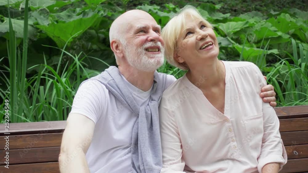 Senior couple sitting on bench in summer park embracing. Active modern life after retirement. Happy family talking enjoying time together in green garden Senior couple sitting on bench in summer park embracing. Active modern life after retirement. Happy family talking enjoying time together in green garden