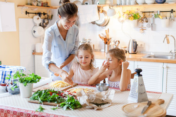 Family are cooking italian pizza together in cozy home kitchen. Cute kids with happy mother are preparing food for dinner and eating cheese. Two girls are helping woman. Lifestyle moment.