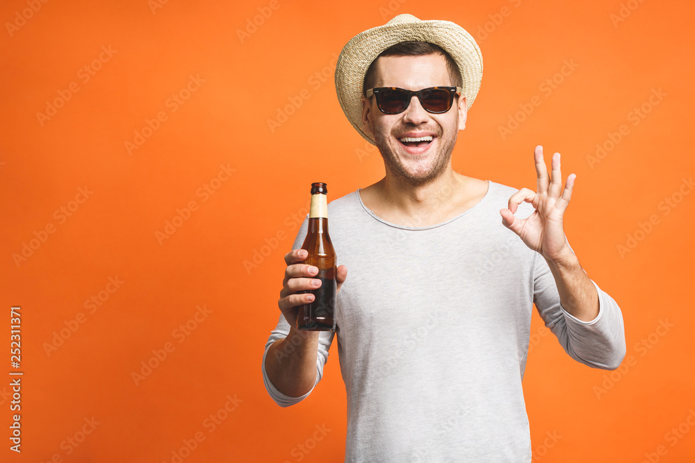 A young guy with a hat and sunglasses isolated on a orange background holds a bottle of beer. Watching football match. Ok sign. A young guy with a hat and sunglasses isolated on a orange background holds a bottle of beer. Watching football match. Ok sign.