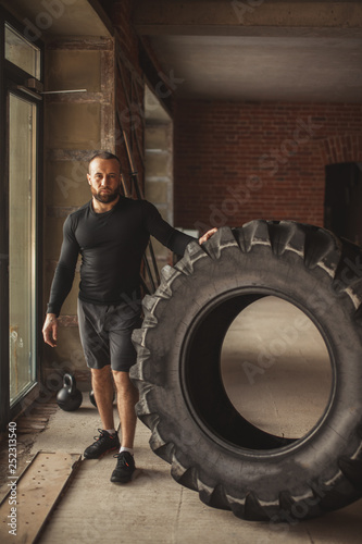 CrossFit Tire Technique. Powerful bodybuilder explains rules and order of training with improvised powerlifting element, standing near huge tire in fitness club